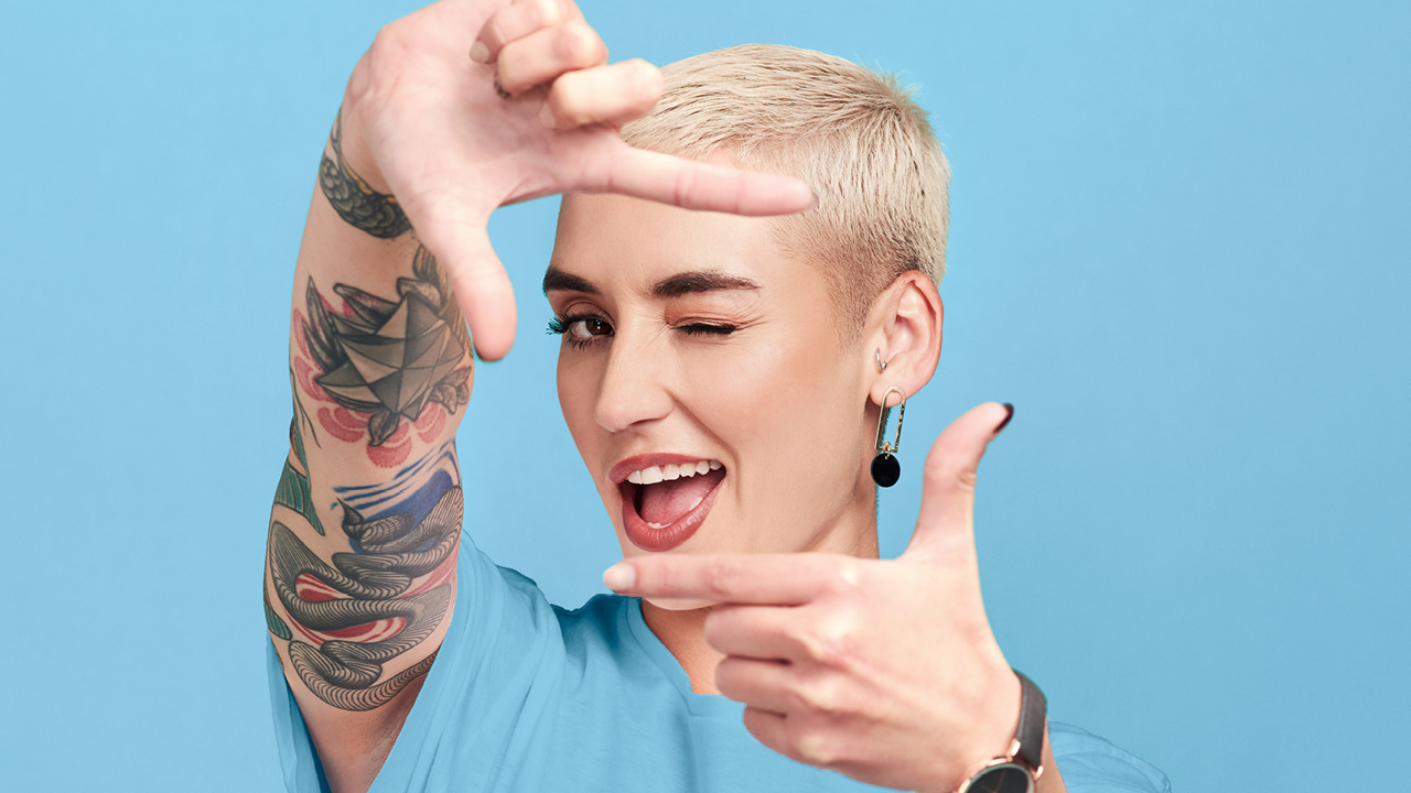 Eine junge Dame mit kurzen, blonden Haaren und Tattoos formt mit ihrem Fingern ein Viereck und blickt zwinkernd hindurch. Sie lächelt dabei verschmitzt.